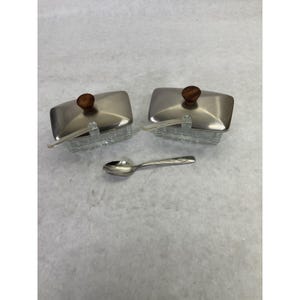 May include: Two rectangular glass condiment containers with clear, textured bases and brushed metal lids. Each lid features a small, round, dark brown wooden knob. Two clear plastic spoons are tucked into the containers, and one silver metal spoon lies between them.