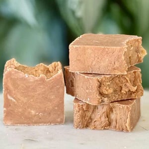 May include: Four light brown, square soap bars are stacked, with rough, textured edges. A single bar, also light brown, stands separately, with a textured, uneven top. The soaps are on a white surface, with a blurred green background.