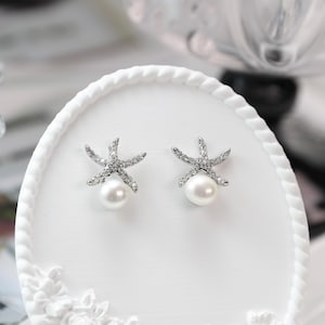 May include: A pair of starfish-shaped stud earrings. Each earring features a white pearl at the base and is adorned with sparkling clear crystals. The earrings are set on a white, decorative surface.