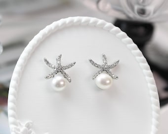 Puede incluir: Un par de pendientes de botón con forma de estrella de mar. Cada pendiente presenta una perla blanca en la base y está adornado con brillantes cristales transparentes. Los pendientes están colocados sobre una superficie blanca decorativa.