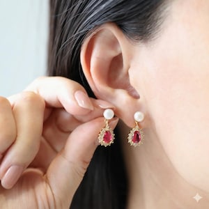 May include: Gold-toned drop earrings featuring a white pearl stud and a teardrop-shaped, ruby-red gemstone. The gemstone is surrounded by a halo of small, clear crystals. The earrings are being worn by a person.