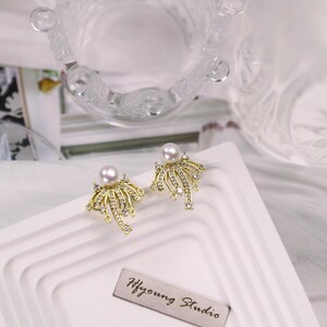 Puede incluir: Un par de pendientes de oro con un diseño de perla y cristal. Los pendientes están sobre una superficie blanca con el texto "Hfyoung Studio" escrito en un pequeño rectángulo plateado.