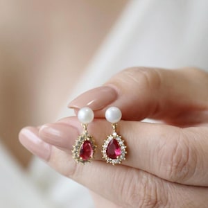 Pode incluir: Um par de brincos dourados com um pino de pérola branca no topo, ligado a uma pedra vermelha em forma de gota pendente, rodeada por um halo de pequenas pedras transparentes. Os brincos são segurados entre os dedos para mostrar os detalhes.