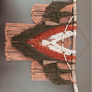 May include: A macrame wall hanging with a geometric design in shades of peach, rust, white, and forest green. The hanging is attached to a natural branch.