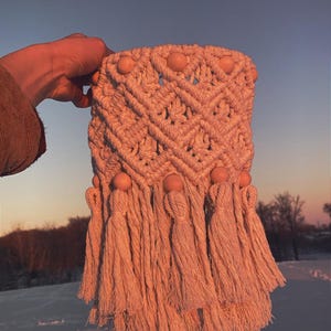 May include: A macrame wall hanging with a geometric pattern and wooden beads. The hanging has a fringe of tassels and is a light beige color.