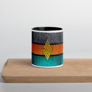 Puede incluir: Una taza de cerámica blanca con un borde y un asa negros. La taza presenta un diseño geométrico con una forma de diamante amarillo en el centro, rodeado de bandas con patrones naranja, azul y gris.