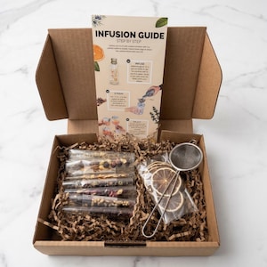 Puede incluir: Una caja de cartón marrón llena de botánicos secos, un colador y una guía de infusión paso a paso. La caja contiene varios tubos de vidrio llenos de coloridos ingredientes secos y un paquete de rodajas de cítricos secos.
