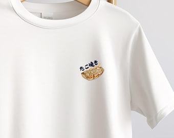 May include: White t-shirt with a small graphic of takoyaki, a popular Japanese street food, in a brown paper boat. The Japanese text "たこ焼き" is above the food illustration. The shirt is displayed on a wooden hanger.