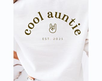 May include: White sweatshirt with the text "cool auntie" in a brown arch with a rock and roll hand symbol in the center. The text "EST. 2025" is below the arch.