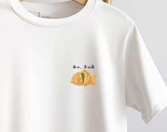 May include: White t-shirt with a small graphic of a fish-shaped pastry with green and tan details, and Japanese text above it. The shirt is hanging on a wooden hanger.