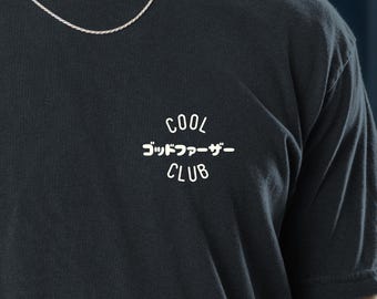 May include: Black t-shirt with the text "COOL ゴッドファーザー CLUB" in white. The shirt has short sleeves and a crew neck. A silver chain necklace is visible at the neckline.