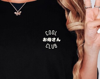May include: Black t-shirt with the words "COOL お母さん CLUB" printed in white. A gold butterfly pendant hangs from a delicate necklace. The image shows a close-up of the shirt and the person's arm making a peace sign.