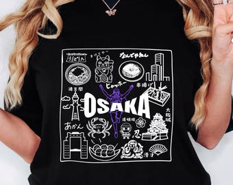 May include: Black t-shirt with a white and purple graphic design featuring the word "OSAKA" and Japanese illustrations. The design includes images of food, landmarks, and text in Japanese characters. A person is wearing the shirt.