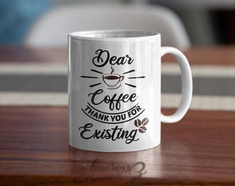 May include: A white ceramic coffee mug with the text "Dear Coffee Thank You For Existing" in black script. The mug features a coffee cup graphic with radiating lines. The mug is on a wooden surface.
