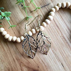 May include: A pair of gold leaf-shaped earrings with delicate cutouts. The earrings are hanging from a wooden surface with a strand of wooden beads.