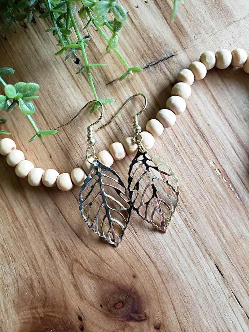 May include: A pair of gold leaf-shaped earrings with delicate cutouts. The earrings are hanging from a wooden surface with a strand of wooden beads.