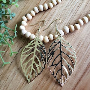 May include: A pair of gold leaf-shaped earrings with a delicate, intricate design. The earrings are hanging from a wooden surface.
