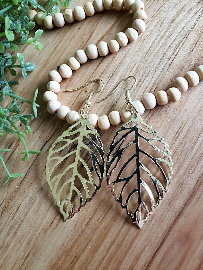 May include: A pair of gold leaf-shaped earrings with a delicate, intricate design. The earrings are hanging from a wooden surface.