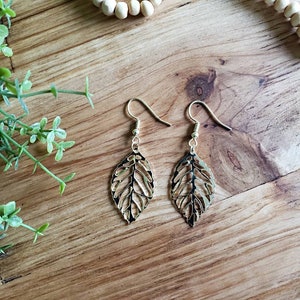 May include: Gold leaf earrings with a delicate, intricate design. The earrings are on a wooden surface.