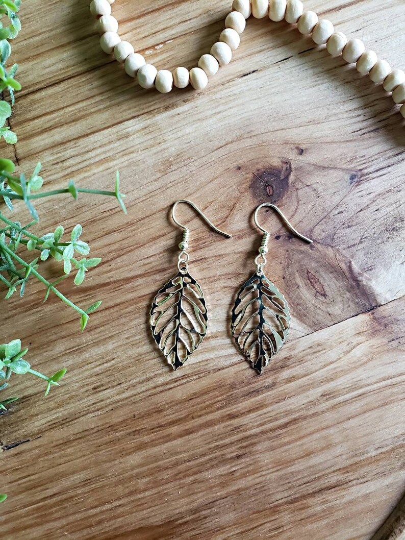 May include: Gold leaf earrings with a delicate, intricate design. The earrings are on a wooden surface.