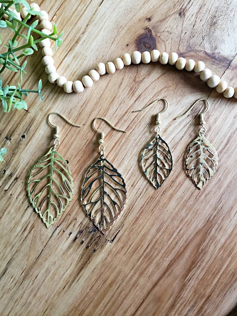 May include: Set of three gold leaf-shaped earrings with delicate cutouts. The earrings are on a wooden surface.