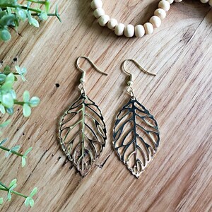 May include: A pair of gold leaf-shaped earrings with a delicate, intricate design. The earrings are hanging from a wooden surface.