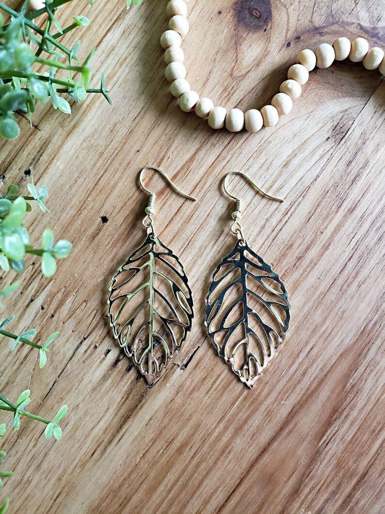 May include: A pair of gold leaf-shaped earrings with a delicate, intricate design. The earrings are hanging from a wooden surface.
