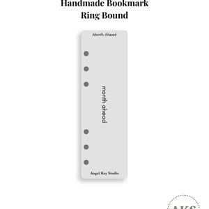 May include: A light grey rectangular ring-bound bookmark featuring six circular punch holes along the left edge. The text "Month Ahead" is printed at the top and vertically along the centre, with "Angel Kay Studio" at the bottom.