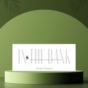 Puede incluir: Una tarjeta rectangular blanca con el texto "IN THE BANK" en gris, un signo de dólar y el nombre "Nozisha Creations" en una fuente cursiva. La tarjeta está sobre un pedestal cilíndrico verde claro contra un fondo verde.