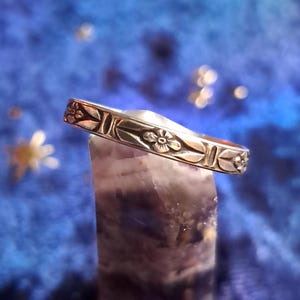 May include: A silver band ring with a floral design. The ring is sitting on a purple and white crystal.