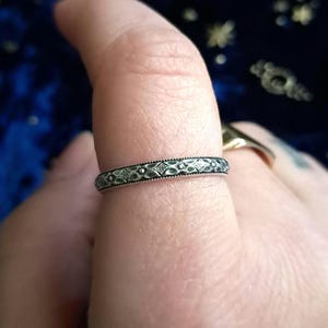 May include: A silver ring with a delicate, patterned design. The ring is worn on a finger.