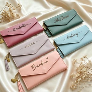May include: Five rectangular envelope-style wallets in pink, mauve, light purple, light blue, and dark green. Each wallet features a gold-tone chain with a matching tassel and heart charm, and is personalized with names: Linabelle, Vivian, Barbie, McKenzie, and Lindsay.
