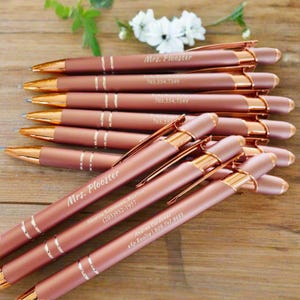 Puede incluir: Un conjunto de bolígrafos de oro rosa con grabados personalizados. Los bolígrafos presentan un acabado en oro rosa y una banda plateada. Los grabados incluyen nombres, títulos e información de contacto.