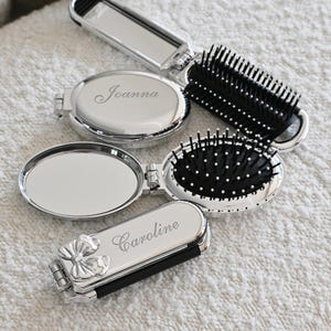 May include: Silver compact mirrors and hairbrushes. Two oval compact mirrors are open, revealing a mirror surface. One mirror is engraved with the name "Joanna", and the other with "Caroline". Two hairbrushes with black bristles are also present.