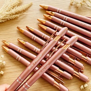 Pode incluir: Uma coleção de canetas esferográficas em ouro rosa fosco com detalhes, clipes e pontas em tom dourado polido. Cada caneta apresenta texto branco personalizado, incluindo nomes como Mrs. Plooster e contatos como 703.554.7349 no corpo.