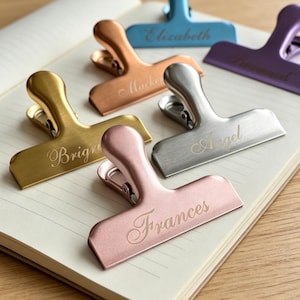 May include: A collection of six metal binder clips in various metallic finishes including gold, rose gold, copper, silver, blue, and purple. Each clip is engraved with a name: Brigne, Frances, Macken, Angel, Elizabeth, and Runaad.