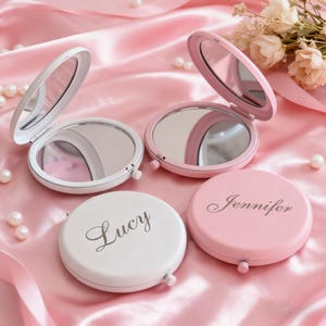 以下が含まれることがあります： 白とピンクのコンパクトミラー。名前「Lucy」と「Jennifer」が刻印されています。一部は開いており、一部は閉じられています。丸い鏡はピンクのサテンの表面に置かれています。背景には真珠と花があります。