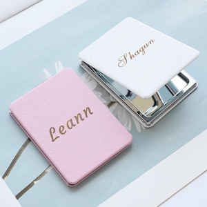 May include: Two personalized compact mirrors. One is white with the name "Shagun" in gold script, the other is pink with the name "Leann" in gold script. Both have a square shape and a silver frame.