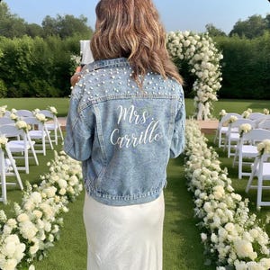May include: Pale blue denim jacket adorned with pearls, featuring the name "Mrs. Carrillo" in white script on the back. The jacket is worn at an outdoor wedding ceremony with floral decorations.