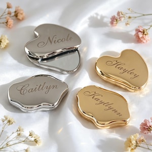 Puede incluir: Cuatro espejos de bolsillo con forma de corazón y acabados metálicos en plata y oro. Cada espejo presenta un nombre grabado en letra cursiva, específicamente Nicole, Hayley, Caitlyn y Kaytlynn, en las tapas lisas y reflectantes.