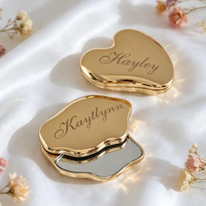 Peut inclure: Deux miroirs de poche dorés en forme de cœur aux bords irréguliers et organiques. Un miroir est fermé et gravé du prénom Hayley en écriture cursive. L'autre miroir est entrouvert, révélant la vitre, avec le prénom Kaytlynn gravé dessus.