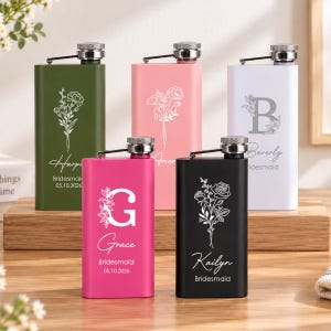 Könnte beinhalten: Fünf rechteckige Metallflachmänner in Olivgrün, Hellrosa, Weiß, Pink und Schwarz. Jeder Flachmann hat einen silbernen Schraubverschluss mit Scharnierarm und ist mit weißen Blumenmotiven, Namen, dem Titel Bridesmaid und dem Datum 05.10.2026 personalisiert.