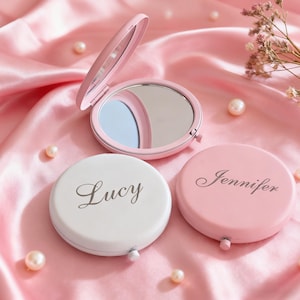 Pode incluir: Três espelhos de bolso redondos com tampas articuladas. Um espelho está aberto, revelando uma superfície refletora. Os outros dois estão fechados, um branco com o nome Lucy gravado em letra cursiva e um rosa com o nome Jennifer.