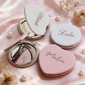 Könnte beinhalten: Eine Sammlung personalisierter Kompaktspiegel in Weiß und Rosa. Ein Spiegel ist herzförmig, die anderen sind rund oder muschelförmig. Jeder Spiegel hat einen Namen in goldener Schrift: Michelle, Lulu und Malia. Die Spiegel liegen auf einer rosa Satin-Oberfläche.