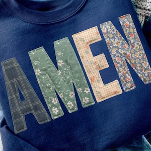 Może przedstawiać: Granatowa bluza z napisem "AMEN" na przodzie, wykonanym z różnych wzorów kwiatowych i kratowych. Bluza jest złożona na parze jasnoniebieskich dżinsów. Widoczna jest również złota bransoletka z łańcuszka i notatnik ze zdjęciami.