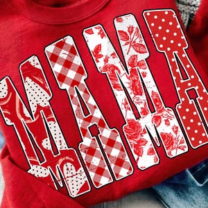 Könnte beinhalten: Rotes Sweatshirt mit dem Wort "MAMA" in großen, weißen Buchstaben. Die Buchstaben sind mit Vichy-Karo, Rosen und Tupfen gemustert. Das Sweatshirt liegt auf einer Jeans, einem Armband und einem Notizbuch.