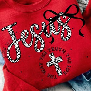 May include: A red sweatshirt with the word "Jesus" in a white, dalmatian-print font. A black bow is above the word. The phrase "The Way, The Truth, The Life" surrounds a cross. The text "John 14:6" is also visible.