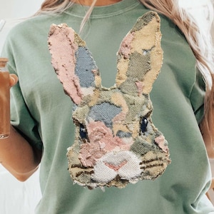 May include: Sage green t-shirt featuring a detailed, textured bunny face design. The bunny's face is composed of various fabric patches in pastel colors, including pink, blue, and yellow. The shirt has a relaxed fit and a crew neck.