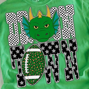 Peut inclure: T-shirt vert avec un motif de dragon et l'inscription "DRAGON TOWN". Le dragon a des accents verts et jaunes. Le ballon de football a un motif léopard. Un jean bleu clair est en arrière-plan. Le texte "digital download" est dans l'image.