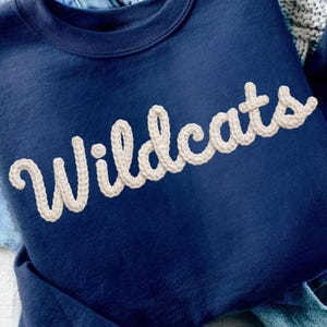 Puede incluir: Una sudadera azul marino con la palabra "Wildcats" en una fuente crema texturizada. La sudadera está colocada sobre un par de vaqueros azul claro. También se ven una pulsera dorada y un cuaderno con fotos.